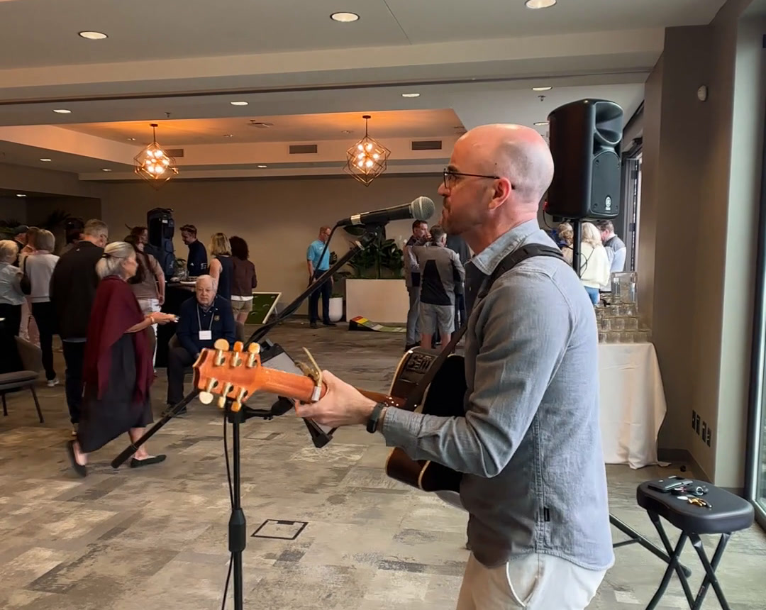 Colin playing acoustic guitar at a mic in a bright function room, filmed from the phone clipped to his speaker stand.