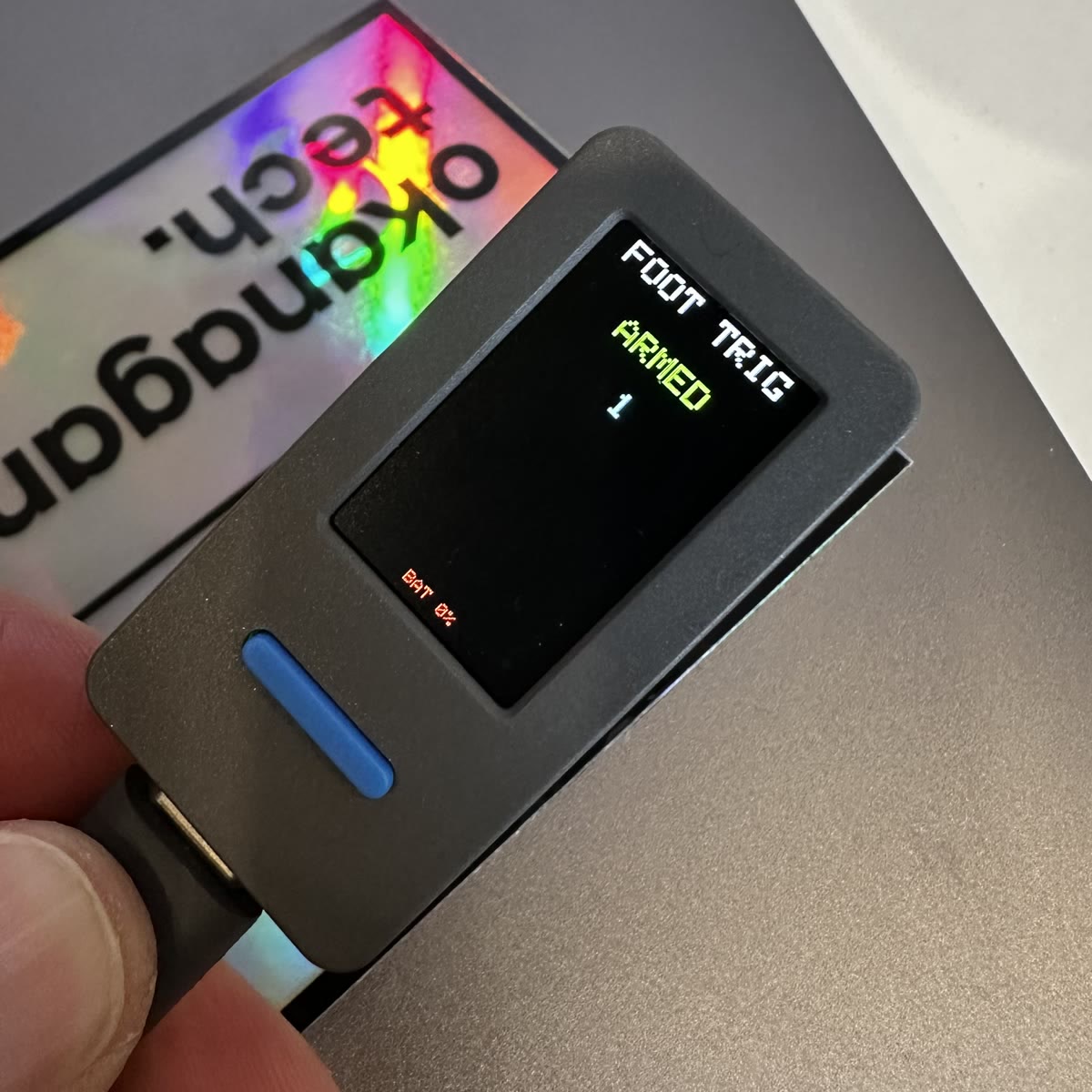 The foot stomper in Colin's fingers, a small grey board with a blue button, its screen reading FOOT TRIG, ARMED in green, tap counter at 1.