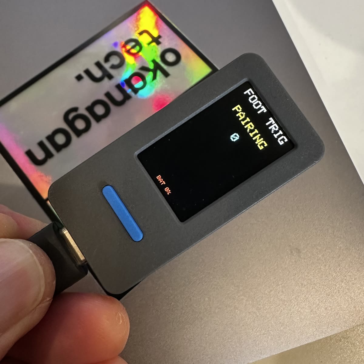 The foot stomper held in Colin's fingers, a small grey M5StickS3 with a blue button, its screen reading FOOT TRIG, PAIRING in yellow, and a tap counter at 0.