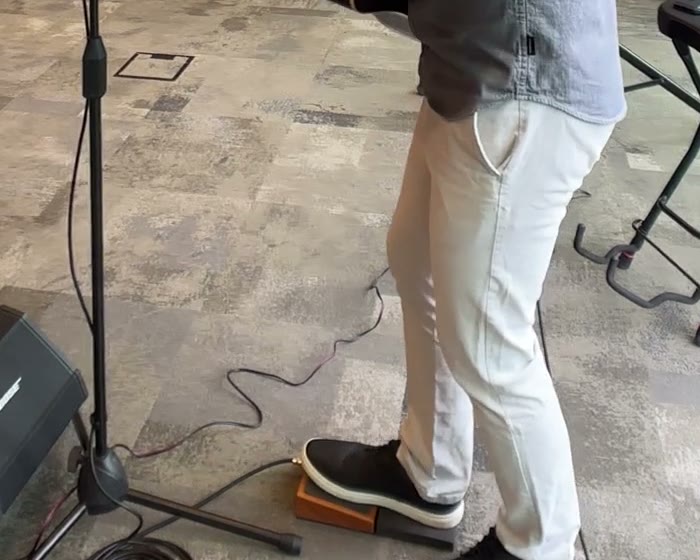 Colin's legs at a gig, right shoe resting on a small wooden stomp box on the carpet, weight on the left leg, mic stand and cables beside him.